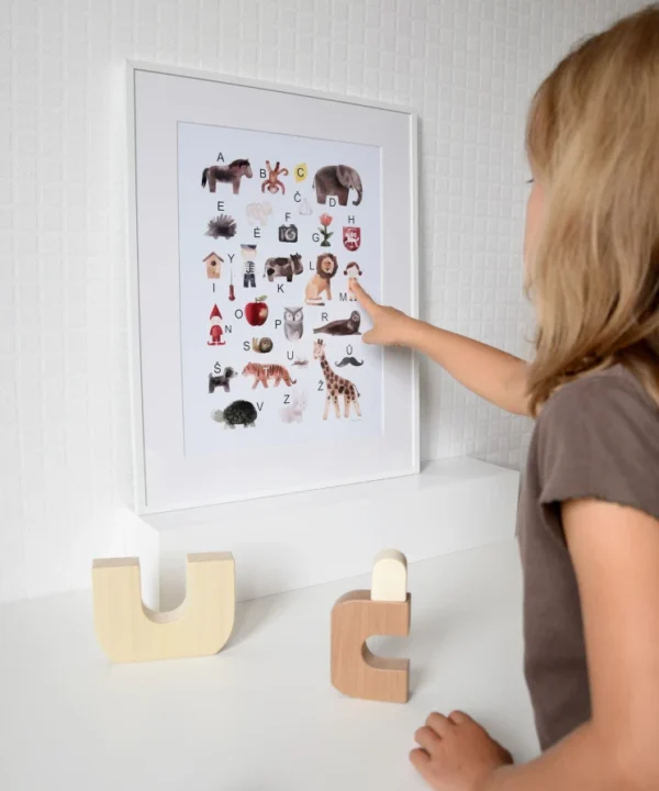 Vaikas rodo į įrėmintą lietuviškos abėcėlės plakatą su iliustracijomis vaikų kambaryje