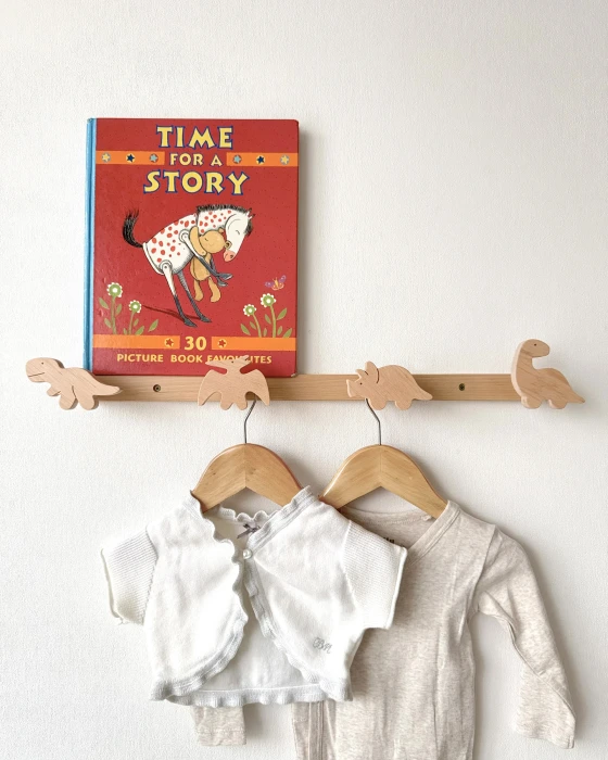 Vaikų kambario medinė kabykla su keturiais dinozaurais, ant kurios atremta vaikiška knyga ir sukabinti kūdikio drabužėliai ant medinių pakabų.