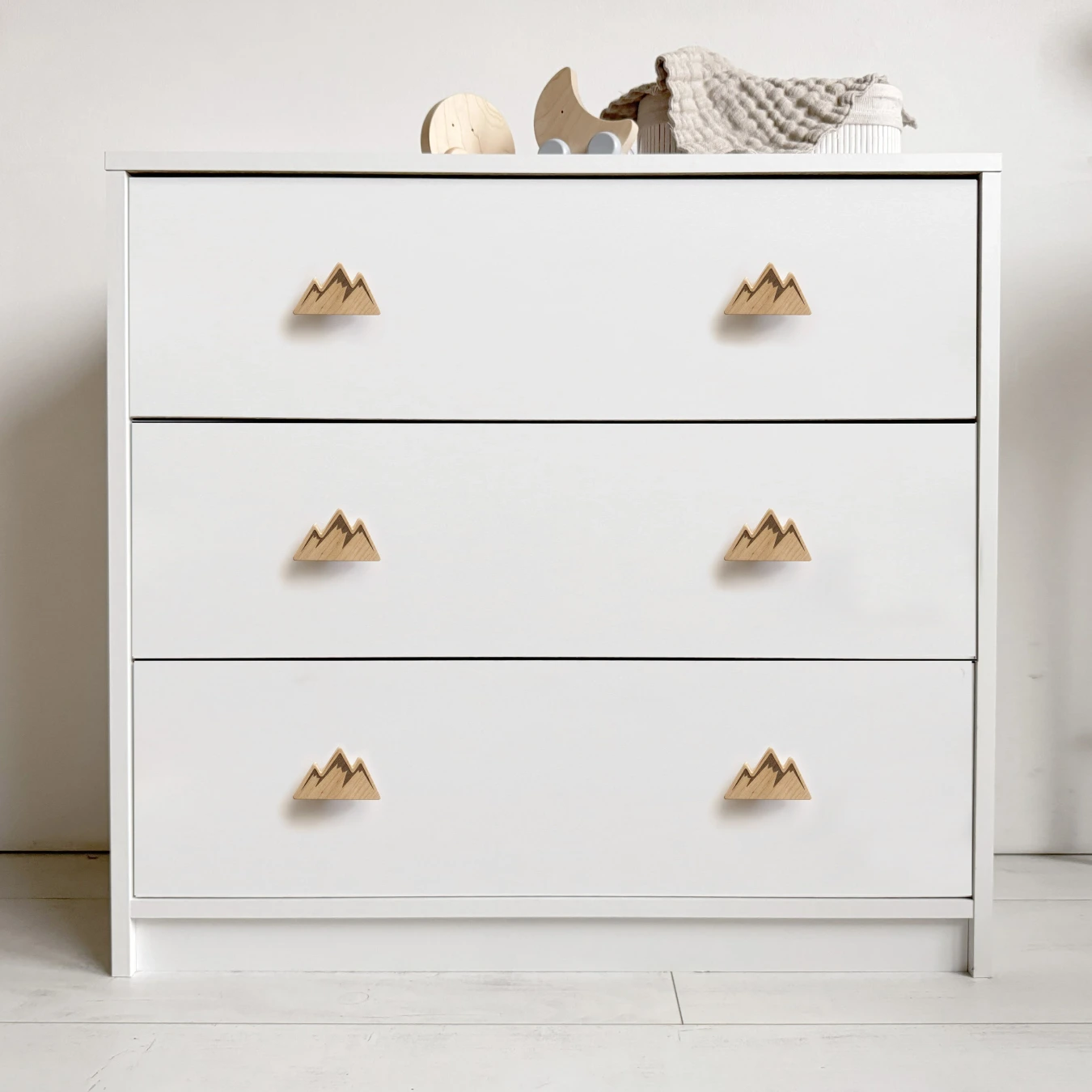 Balta trijų stalčių komoda vaikų kambaryje su šešiomis kalnų formos medinėmis rankenėlėmis.