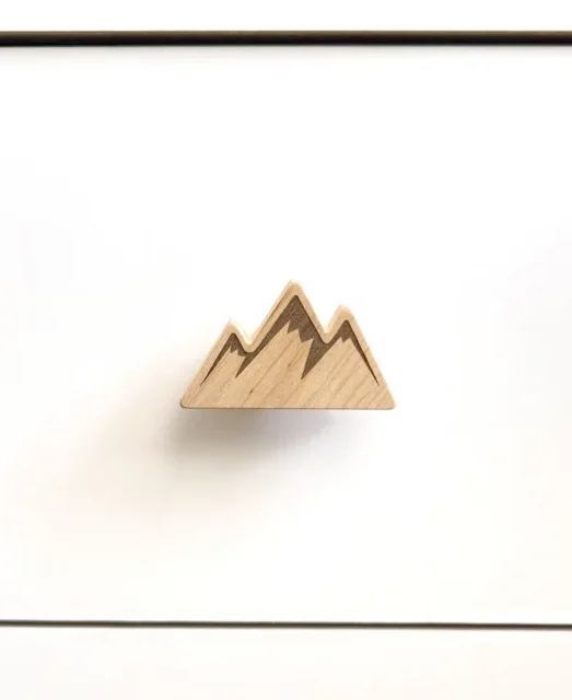 Kalnų formos medinė baldų rankenėlė su graviruotomis viršūnėmis ant balto stalčiaus priekio.