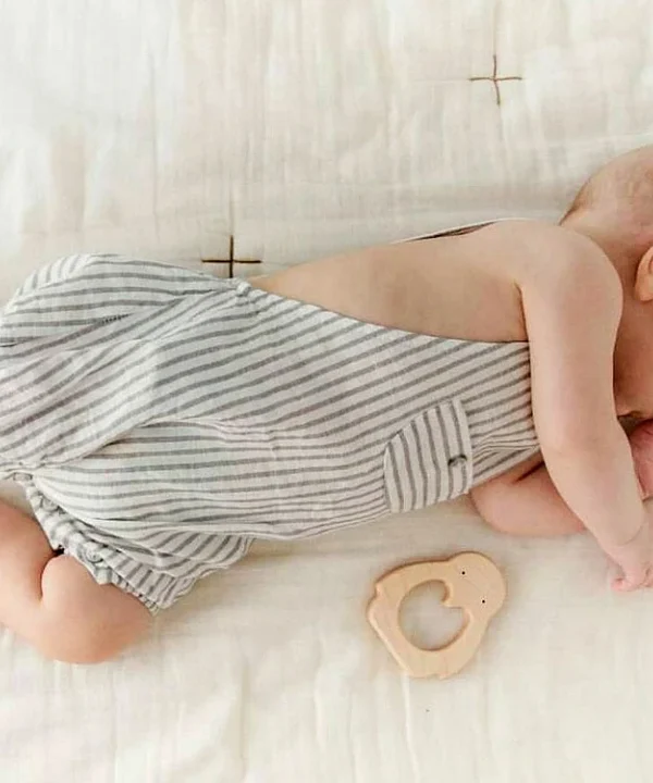 Ant šviesaus pledo gulintis kūdikis su dryžuotu kostiumėliu, šalia kurio padėtas natūralaus medžio kramtukas „Pingvinas“