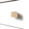 Medinė fotoaparato formos baldų rankenėlė, sumontuota ant baltos minimalistinės komodos stalčiaus šoniniu kampu.