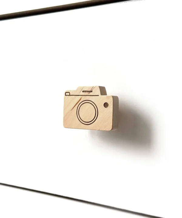 Medinė fotoaparato formos baldų rankenėlė, sumontuota ant baltos minimalistinės komodos stalčiaus šoniniu kampu.