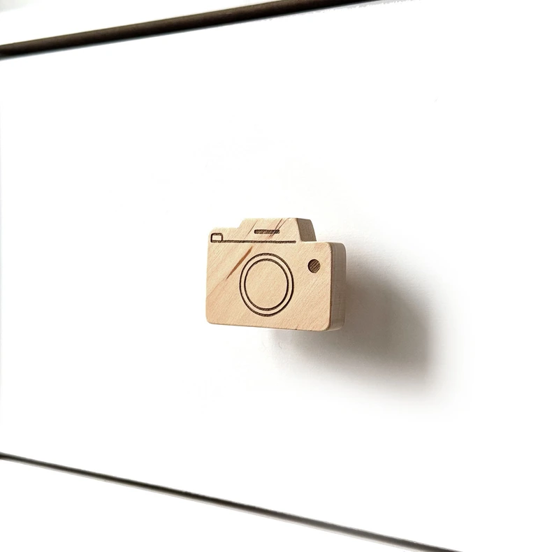 Medinė fotoaparato formos baldų rankenėlė, sumontuota ant baltos minimalistinės komodos stalčiaus šoniniu kampu.