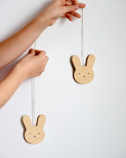 Rankose laikomi du minimalistiniai mediniai Velykų papuošimai zuikučiai su virvutėmis, pagaminti iš šviesaus klevo medžio.