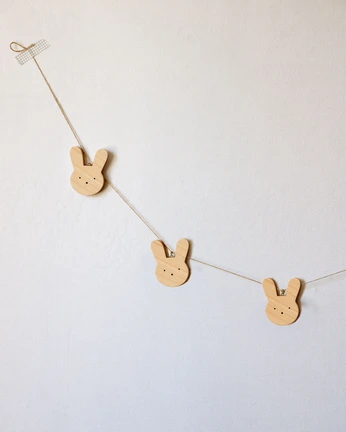 Trijų medinių zuikučių girlianda, priklijuota prie sienos tekstūrine juostele. Matoma natūrali klevo medienos tekstūra ir subtilus minimalistinis dizainas.